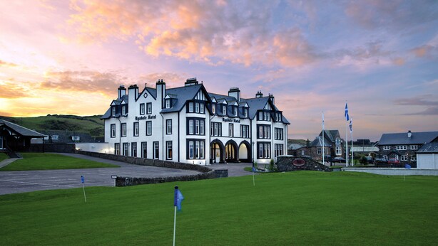A large, white building with multiple chimneys and arched windows, likely a hotel, with a well-maintained lawn and a flag in the foreground. - The Ugadale Hotel and Cottages (Campbeltown)