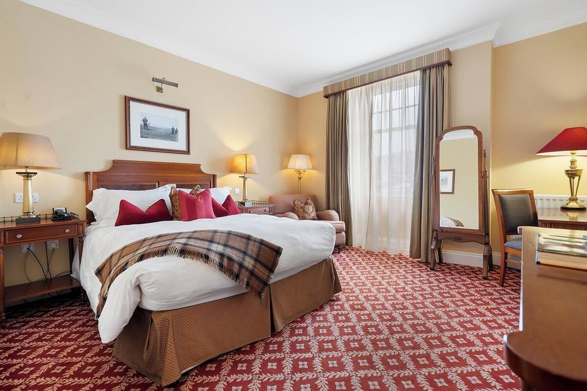 Habitación de hotel con una cama grande, dos mesitas de noche con lámparas, una silla, un escritorio con lámpara, un espejo grande y una ventana con cortinas.