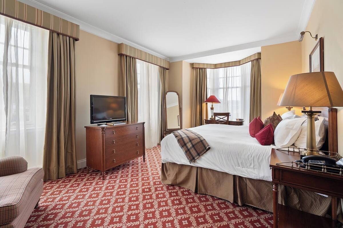 Habitación de hotel con una cama grande, un televisor sobre un aparador de madera, un escritorio con lámpara y una silla.