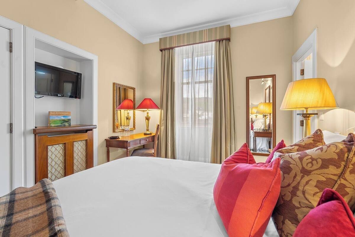Habitación de hotel con cama, escritorio con dos lámparas, televisor, espejo y ventana con cortinas.