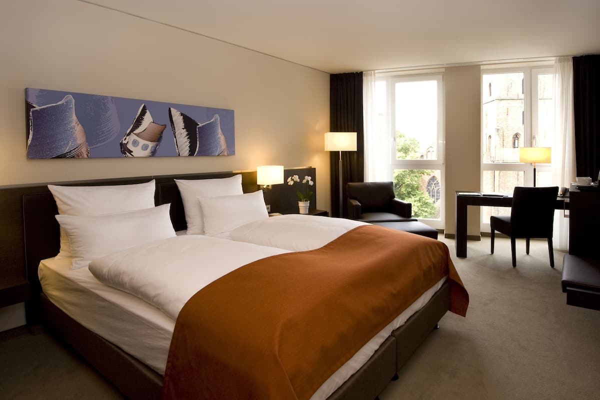 Une chambre d’hôtel avec un grand lit, un bureau, une chaise et une fenêtre donnant sur des immeubles.