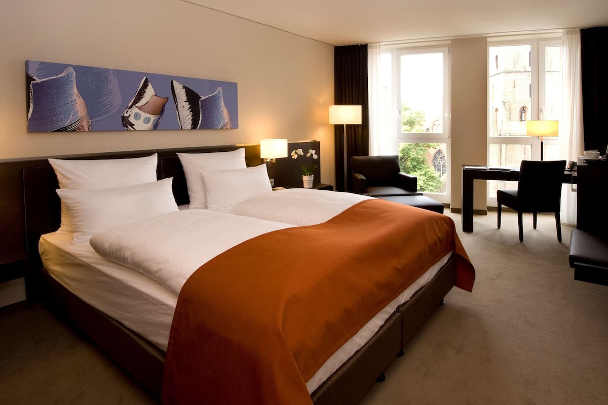 Une chambre d’hôtel avec un grand lit, un bureau, une chaise et une fenêtre donnant sur des immeubles.