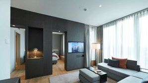 A modern hotel room with a large window, a flat-screen TV, a sofa, and a desk with a lamp.