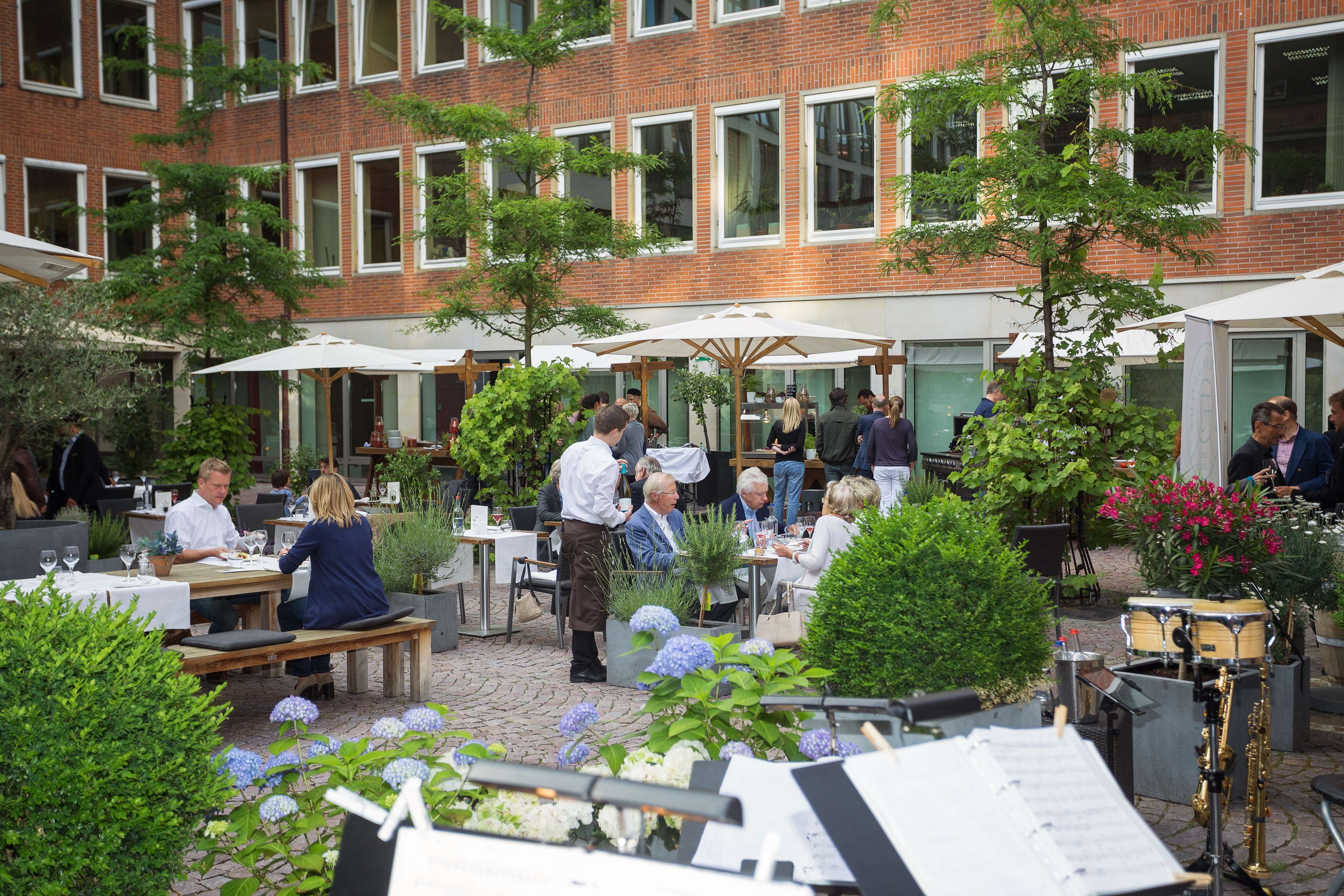 An outdoor dining area with tables and umbrellas, surrounded by greenery and potted plants.