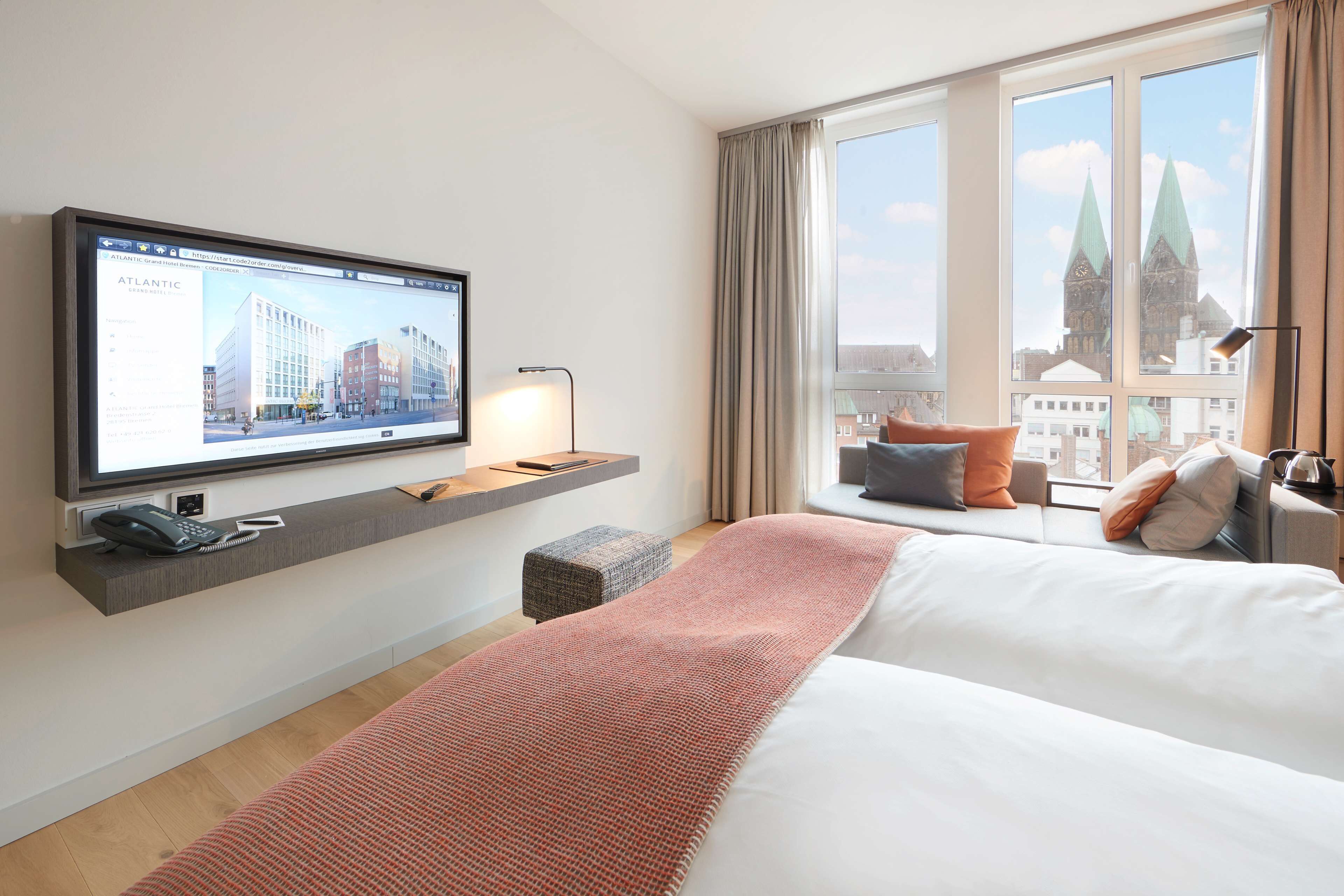 A hotel room with a large window, a flat-screen TV mounted on the wall, a bed with a red blanket, and a small table with a lamp.