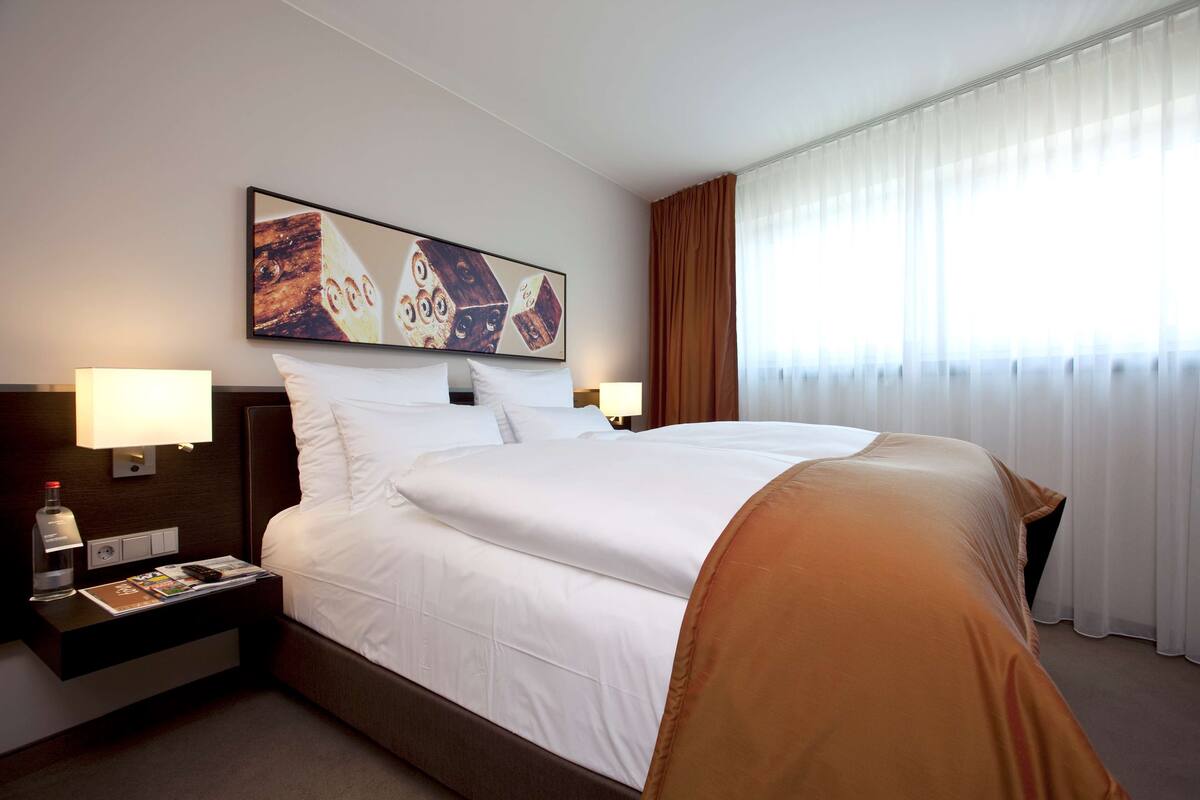 Une chambre d’hôtel avec un grand lit, deux tables de chevet avec des lampes, une petite table avec une bouteille et un magazine, et un tableau accroché au mur.