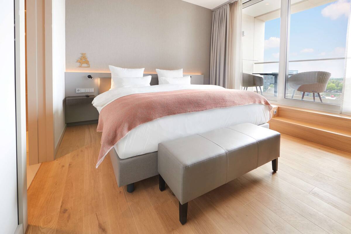 Une chambre moderne avec un grand lit, un banc, une table de chevet et une vue sur la ville grâce à de grandes fenêtres.