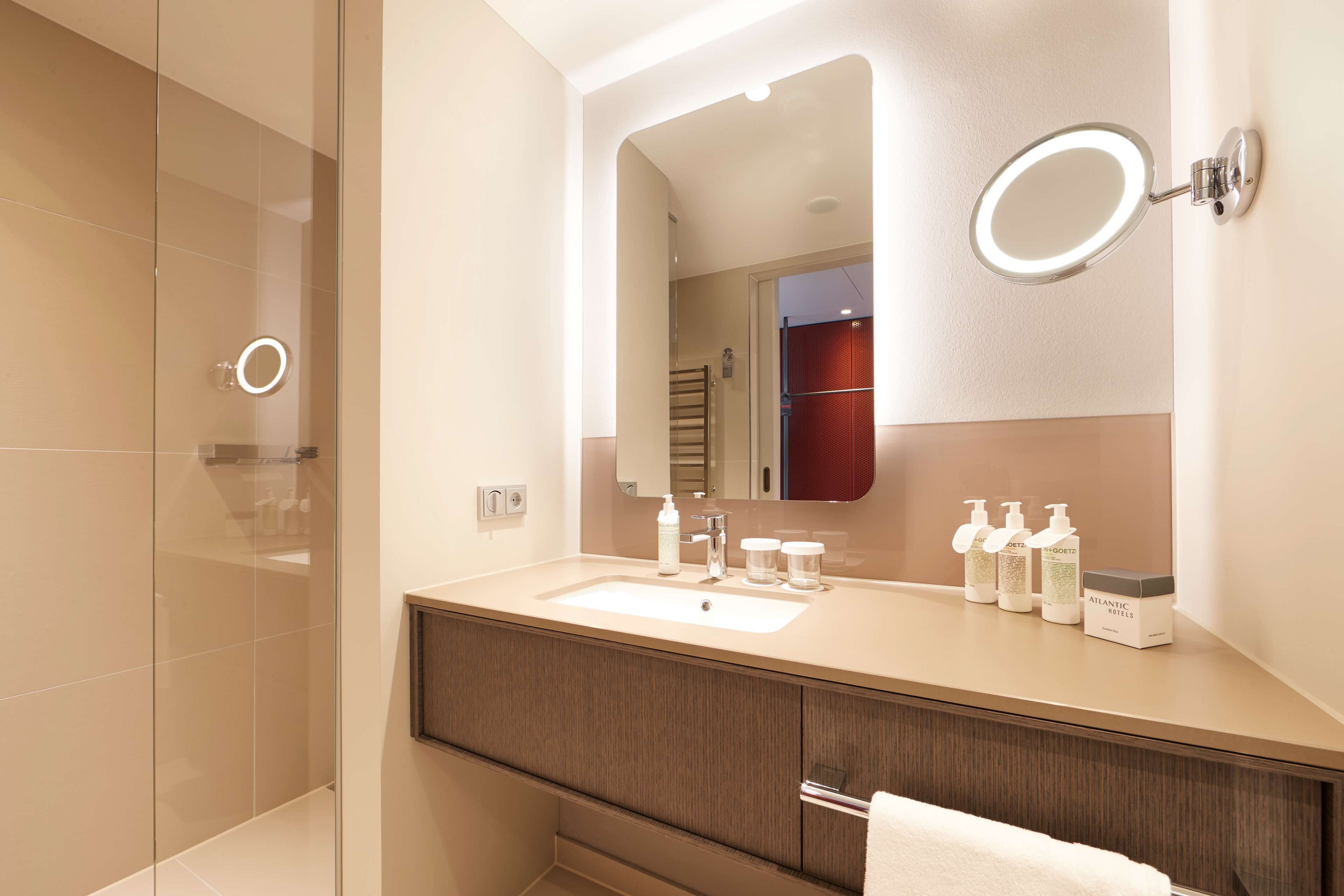 A modern bathroom with a large mirror, a round vanity mirror, and a sink. There are toiletries on the sink counter.