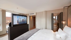 A modern hotel room with a large bed, a flat-screen TV mounted on a dark console, a walk-in closet, and a view of the cityscape through the window.