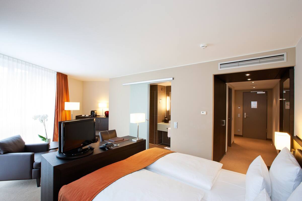 Une chambre d’hôtel avec un lit, un bureau avec une télévision, une chaise, une lampe et une salle de bain visible à travers une porte ouverte.