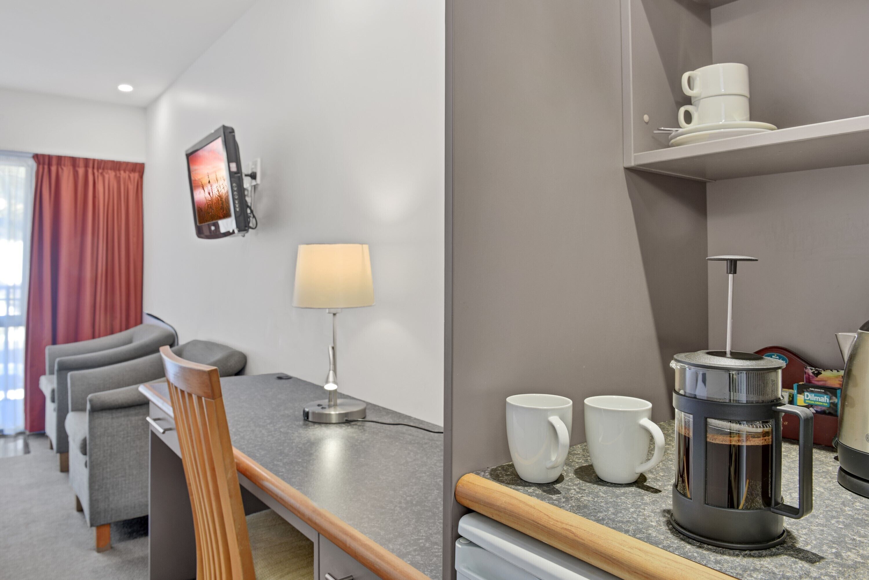 Ein modernes Hotelzimmer mit Essbereich, einem an der Wand befestigten Fernseher, einer Kaffeemaschine und einem Regal mit Tassen.