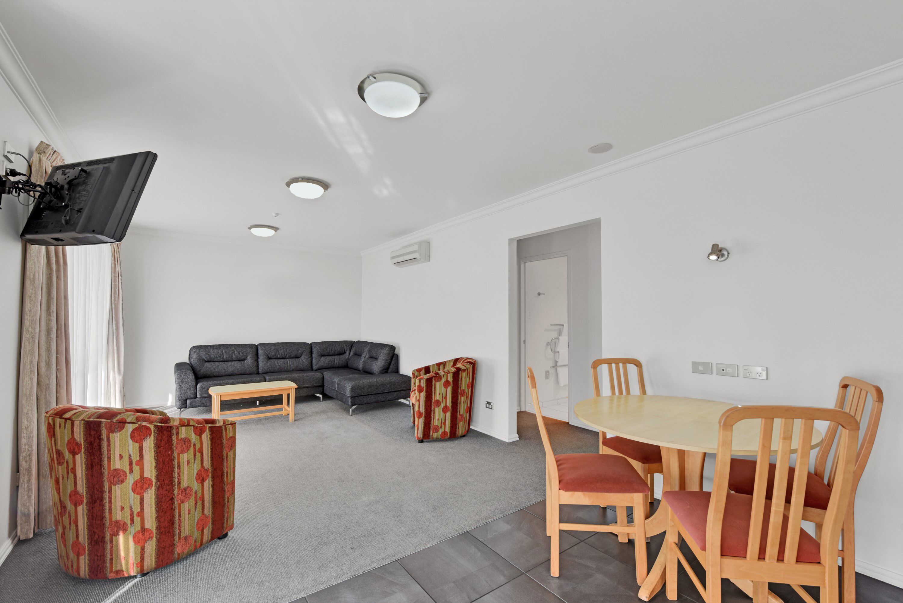Una sala de estar moderna con un sofá gris, una mesa de comedor de madera con cuatro sillas y un reposapiés rojo con estampado.