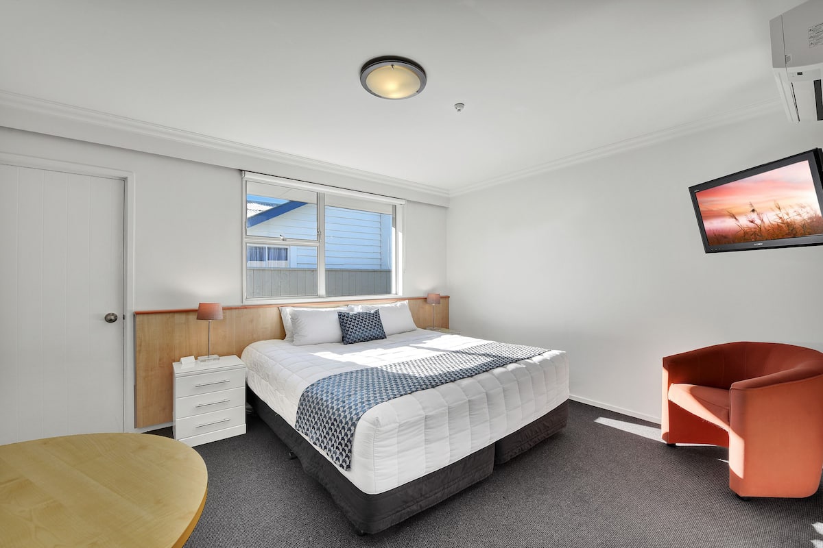 A modern hotel room with a large bed, a desk, a chair, and a TV.