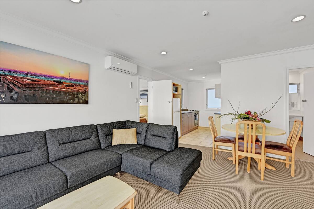 A modern living room with a gray sectional sofa, a wooden dining table with chairs, and a kitchen in the background.