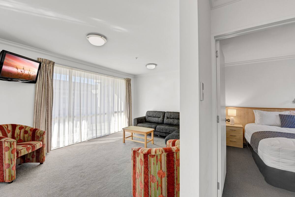 A hotel room with a sofa, a small table, a bed, and a TV mounted on the wall.