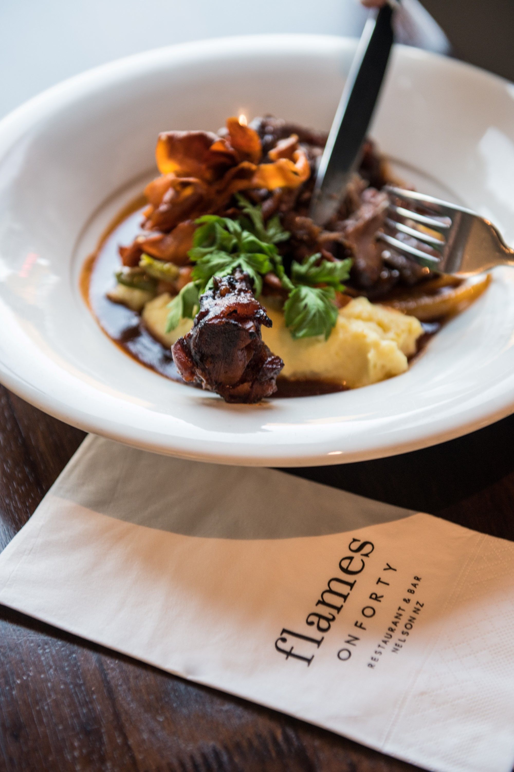 A white plate with a steak, mashed potatoes, and vegetables, accompanied by a fork. A napkin with the text "flames on forty" is visible.