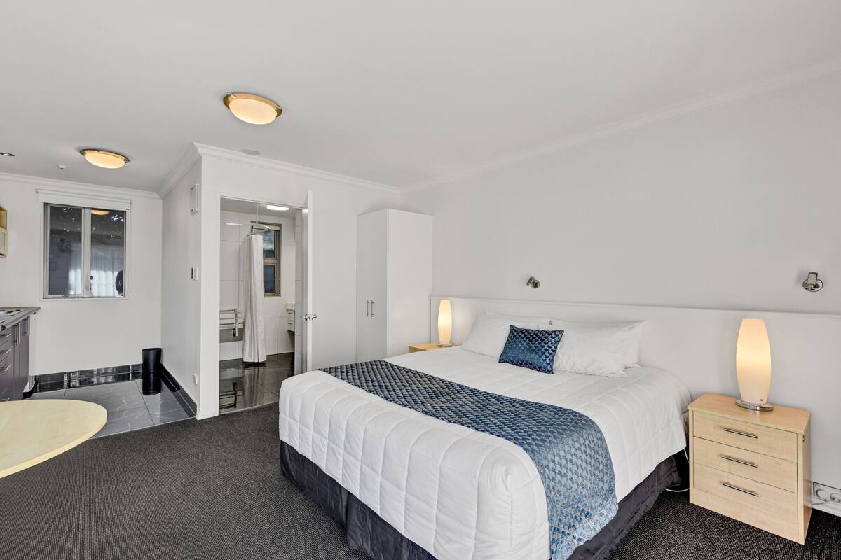 A hotel room with a large bed, bedside lamps, a wooden nightstand, a desk, and a bathroom visible through an open door.