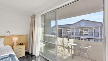Habitación de hotel con cama, mesita de noche, lámpara y puerta corrediza de vidrio que da a un balcón con mesa y sillas.