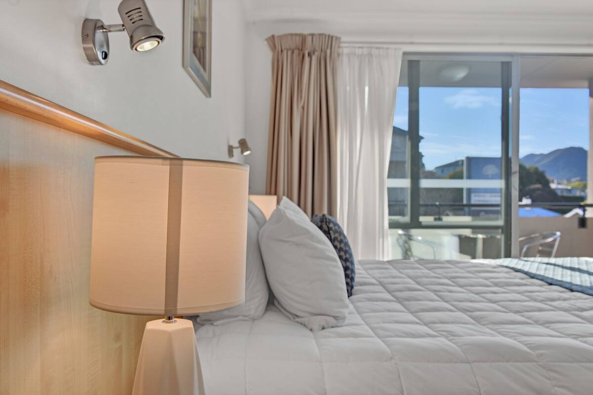 A hotel room with a bed, a lamp, a window with curtains, and a view of the outside.