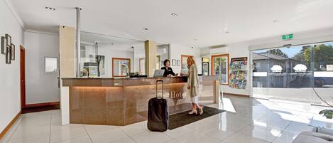 Zona de recepción de hotel con un separador de vidrio, mostrador de recepción, un espejo grande y vistas al exterior.