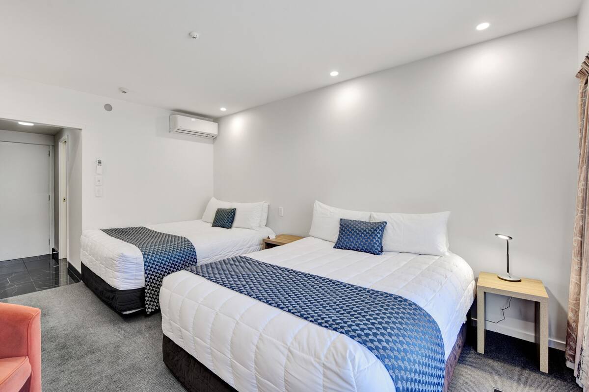A hotel room with two beds, a desk, a chair, and a nightstand with a lamp.
