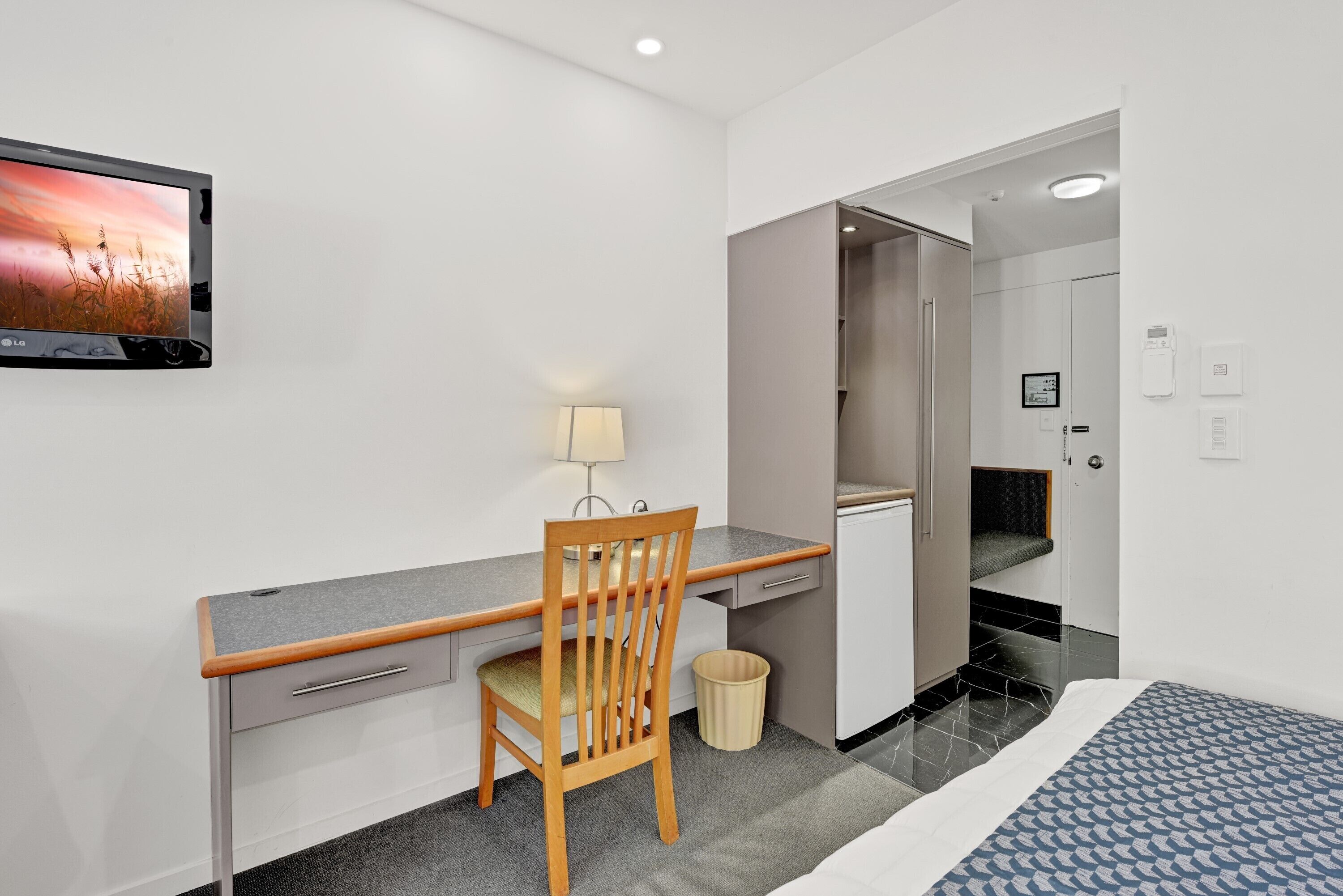 A hotel room with a desk, chair, TV, lamp, and a bed.