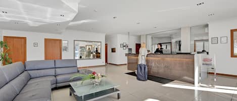 Un hall d’hôtel moderne avec un comptoir d’accueil, un grand canapé d’angle gris, une table basse en verre et une plante en pot.