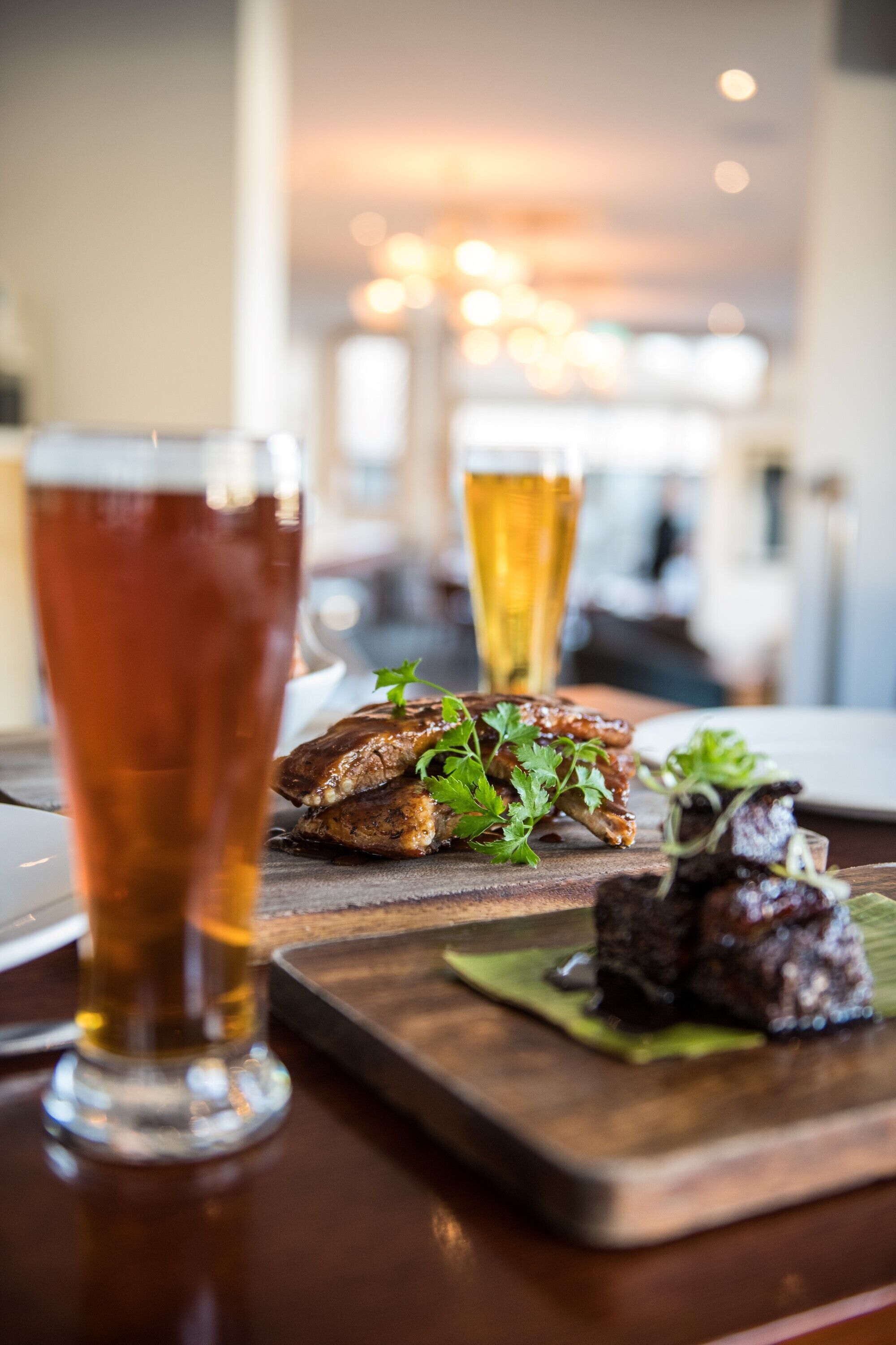 A dining table with a glass of amber-colored beer, a wooden board with grilled meat and greens, and a glass of pale beer.