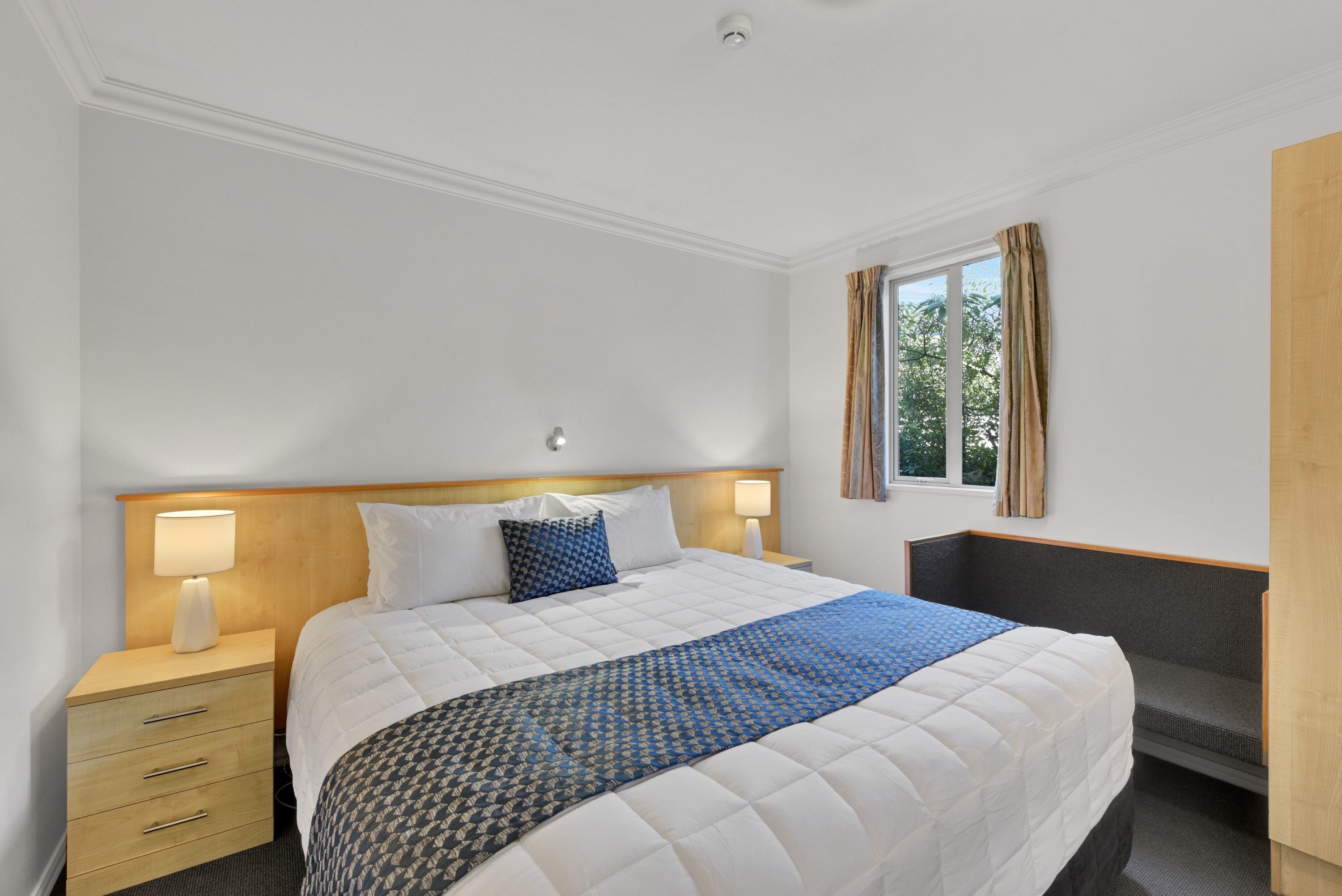 Habitación de hotel con una cama grande, dos mesitas de noche, una ventana con cortinas y un pequeño rincón de descanso.