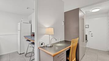 Una cocina compacta con refrigerador, un taburete pequeño, un escritorio con lámpara y una silla.