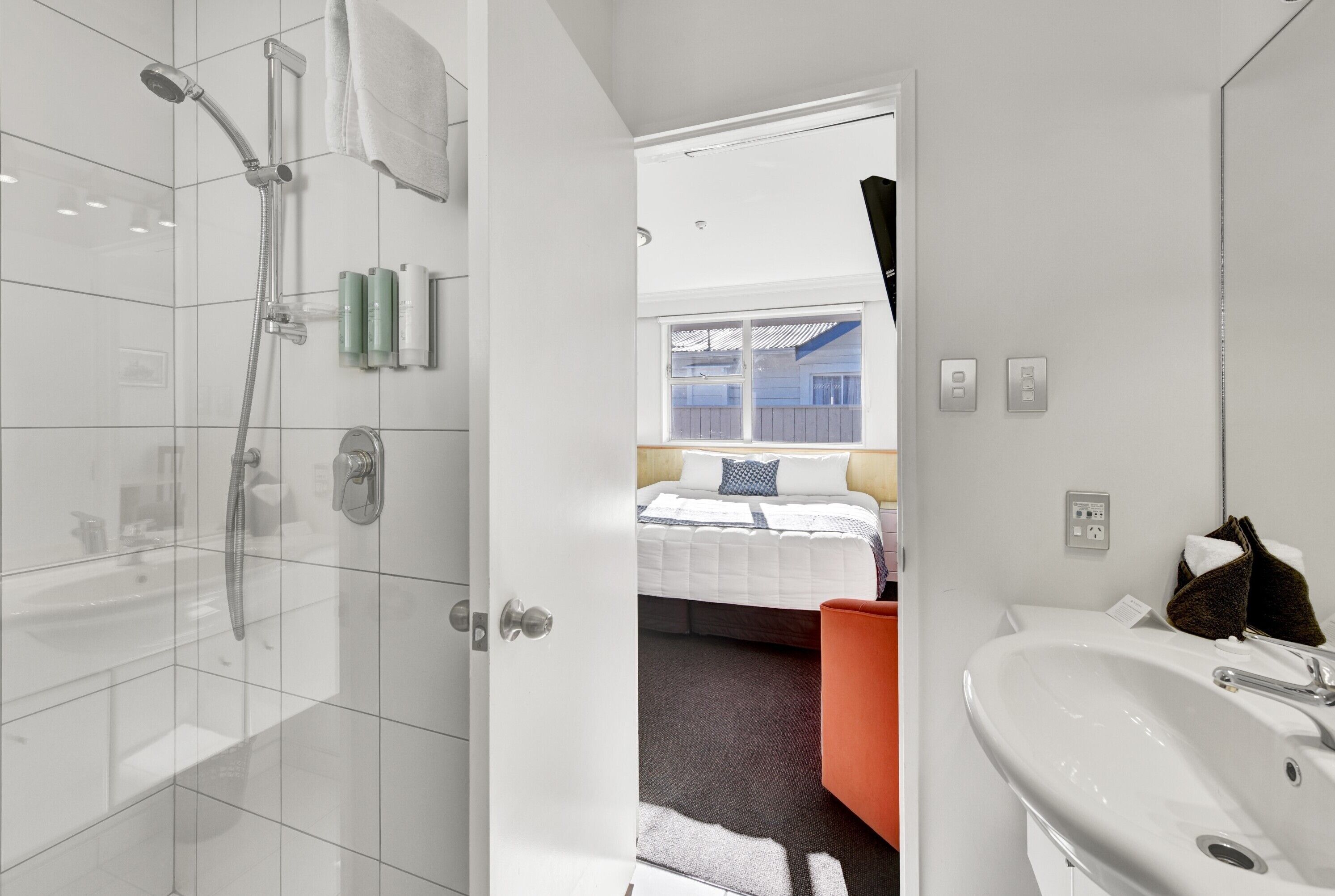 Une salle de bain moderne équipée d’une douche, d’un lavabo et offrant une vue sur une chambre avec un lit et une fenêtre.