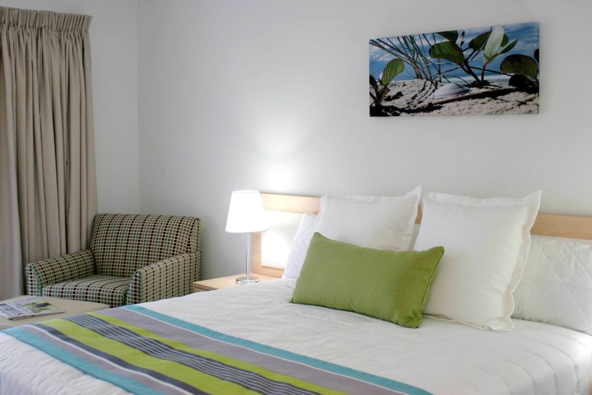 Ein Hotelzimmer mit Bett, Sessel, Nachttisch, Lampe und einem Wandbild.