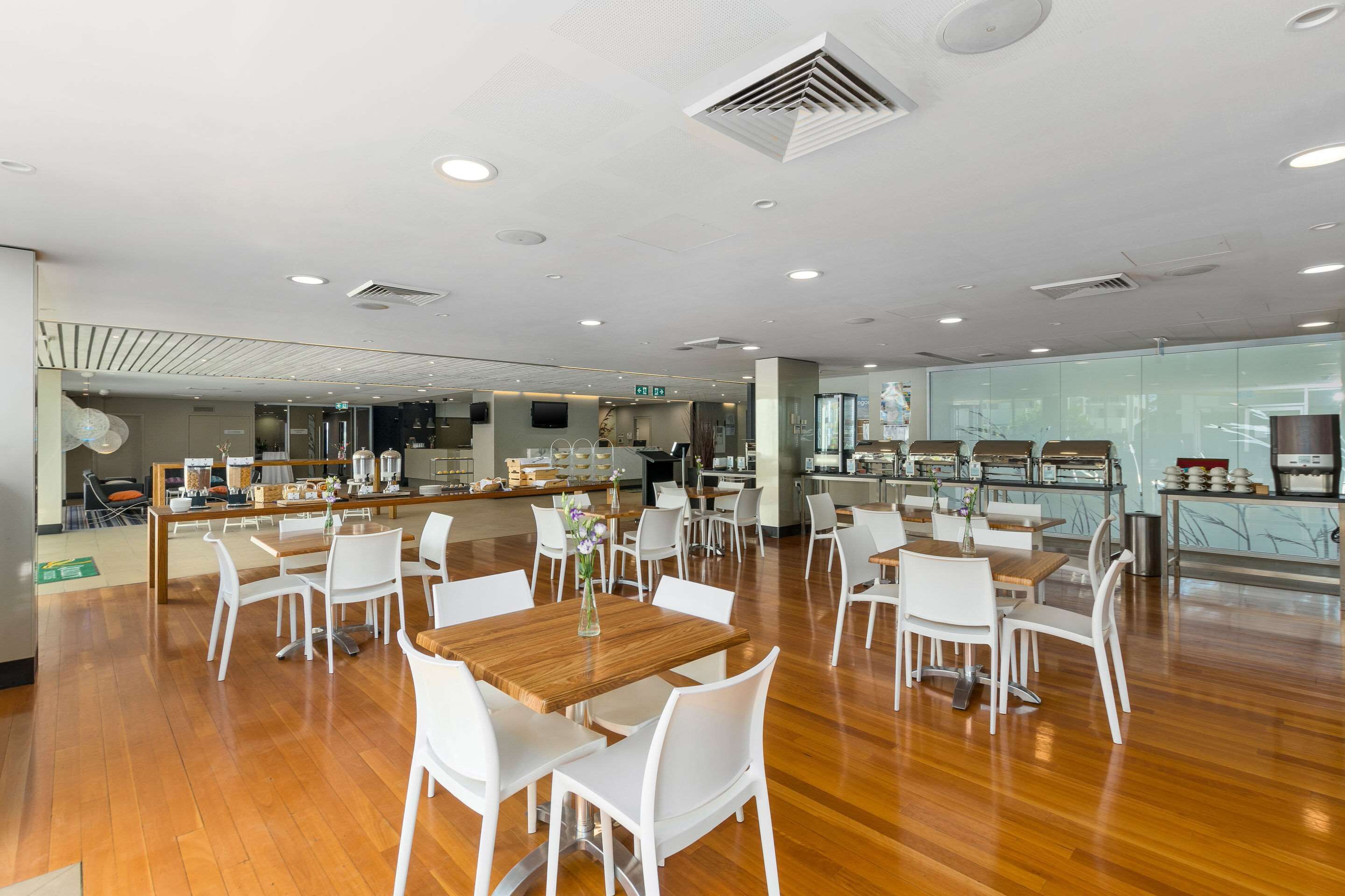 Un comedor amplio con mesas de madera y sillas blancas, una cocina al fondo y un diseño de techo moderno.