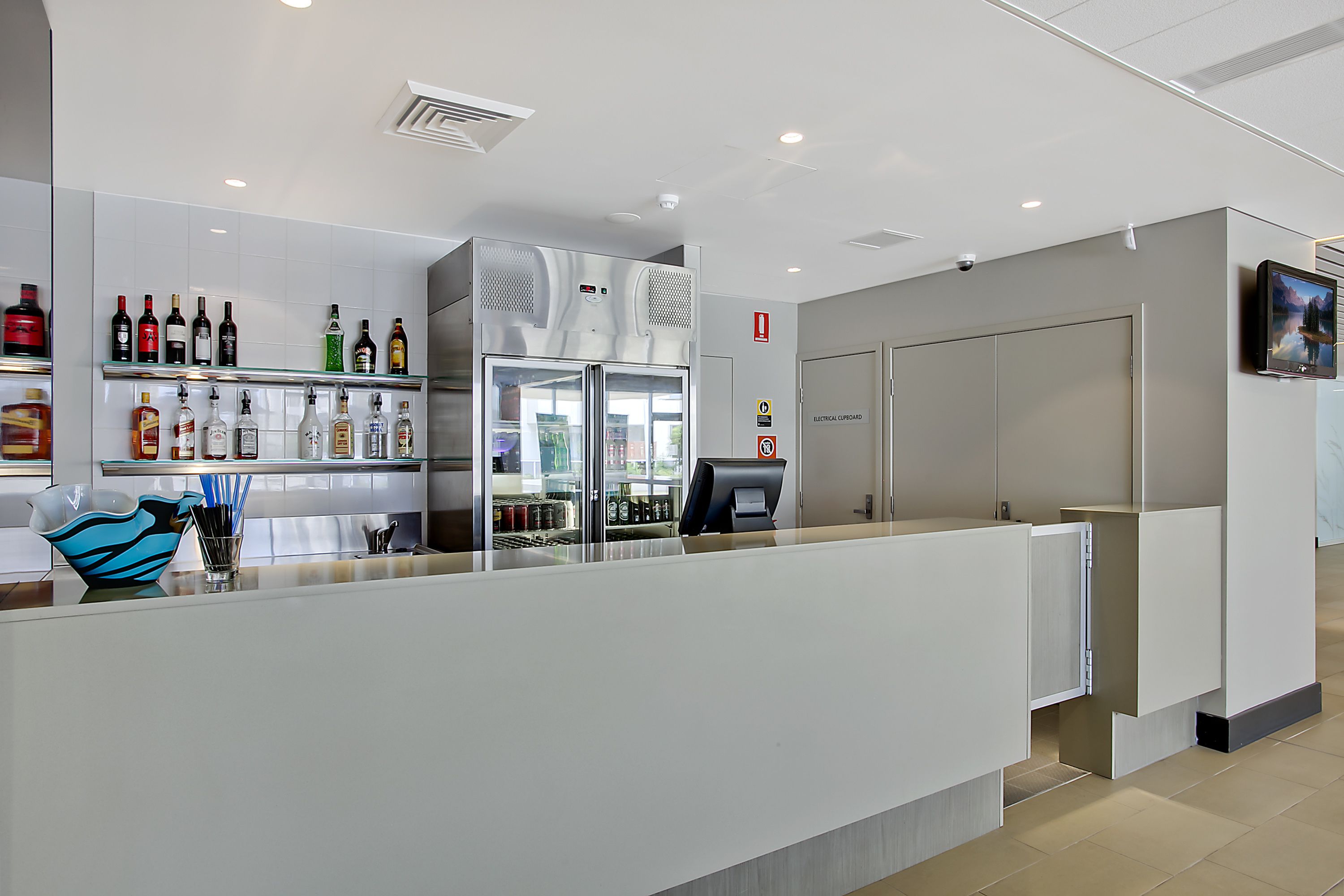 A modern hotel reception area with a glass display of various liquor bottles, a large air conditioning unit, and a closed door to the right.