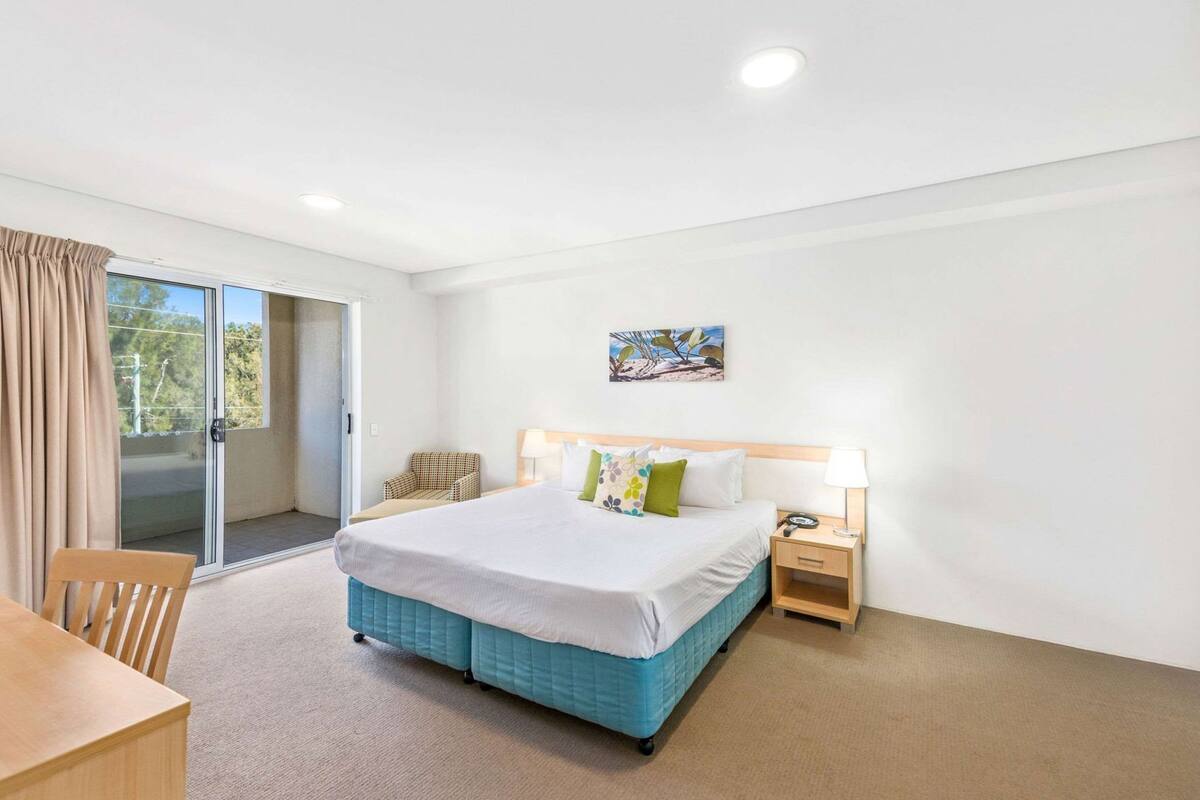 Ein Hotelzimmer mit Bett, Sessel, Schreibtisch, Nachttisch und einem Gemälde an der Wand.