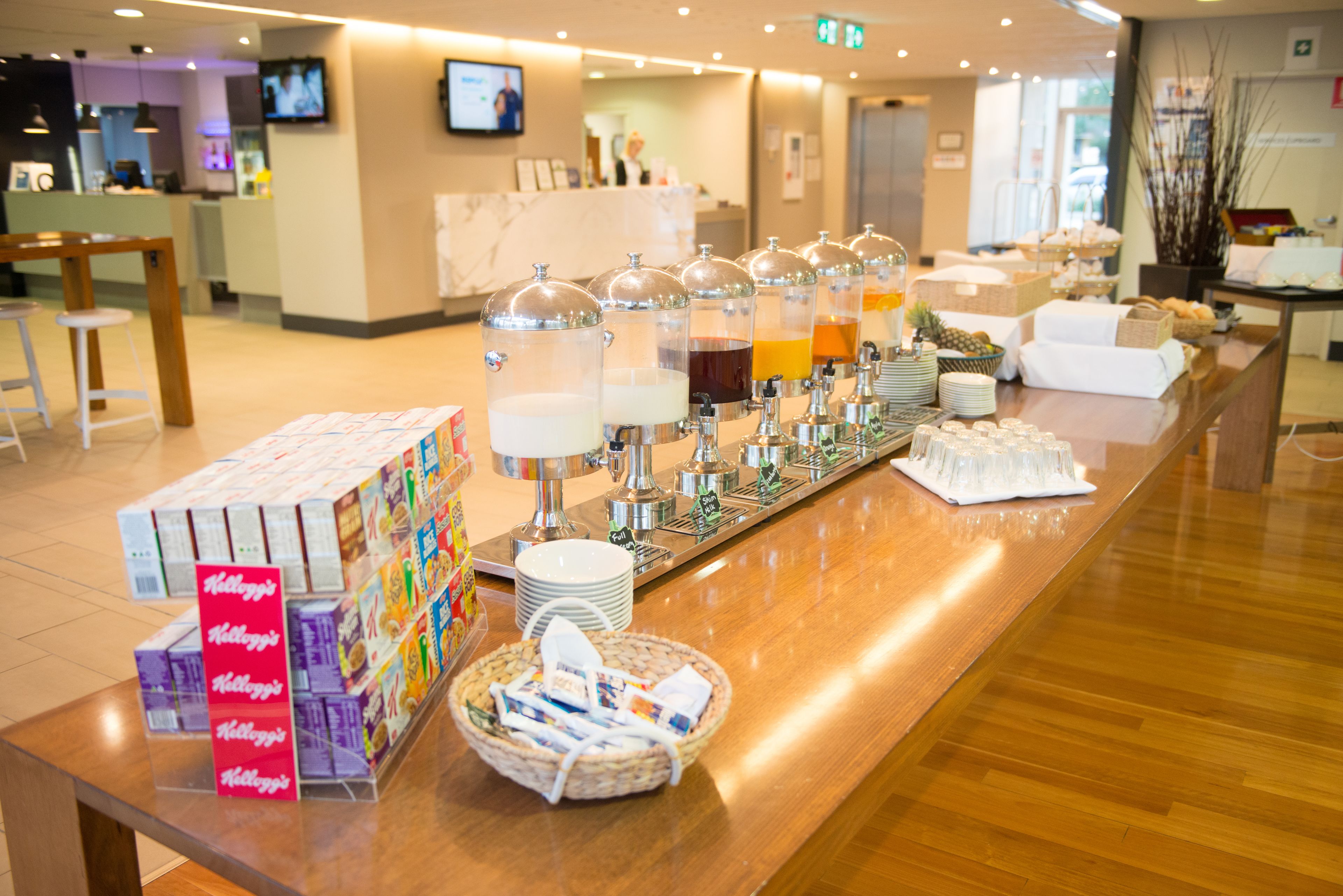 A buffet setup with various food and drink dispensers, a stack of cereal boxes, and a wicker basket.