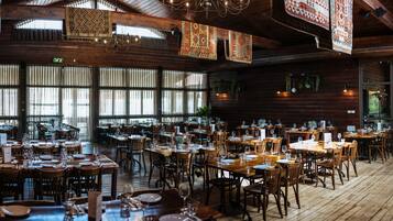 A spacious restaurant with wooden tables and chairs, large windows, and hanging patterned rugs.