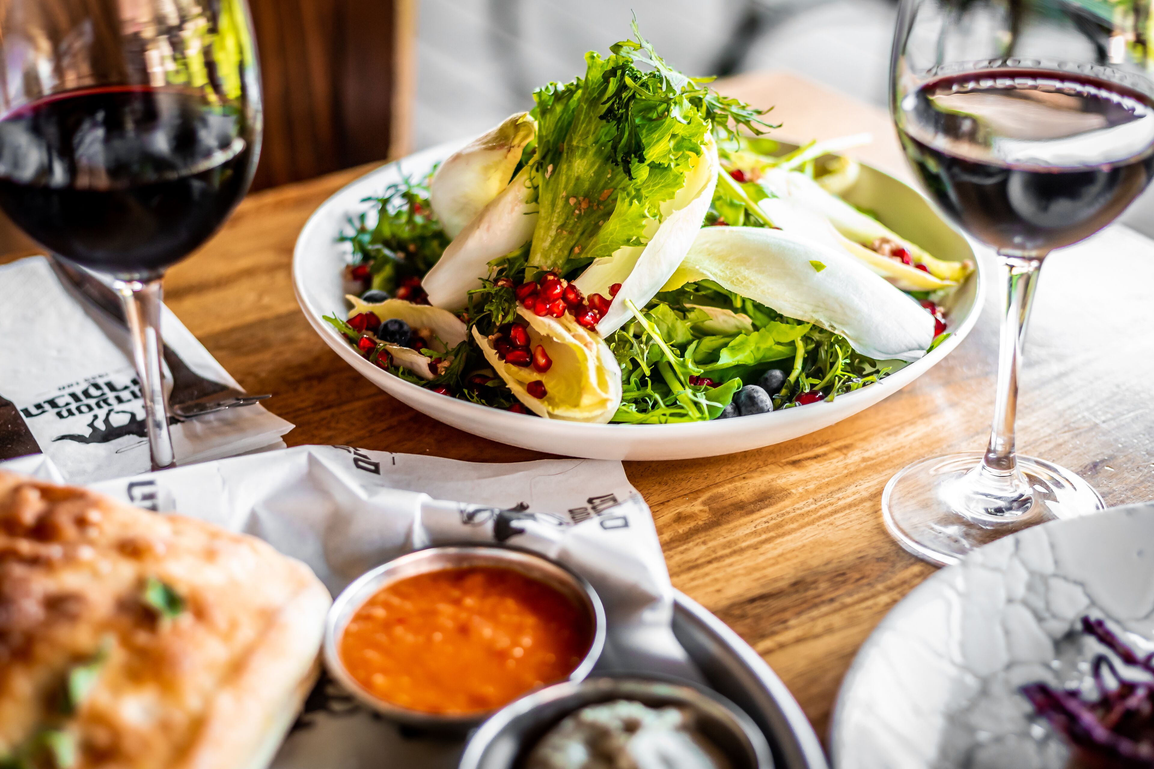 A plate of salad with various greens and pomegranate seeds, accompanied by a glass of red wine and a small bowl of sauce.
