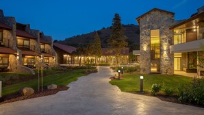 A pathway leading to a stone building with multiple windows and a balcony, surrounded by greenery and a hilly landscape.