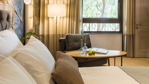 A hotel room with a bed, a round table with a book and apples, a lamp, and a view of trees through a window.