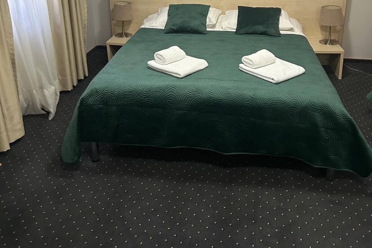 A neatly made bed with green bedding, two pillows, and two rolled towels. There is a wall decoration above the bed and a lamp on each side.