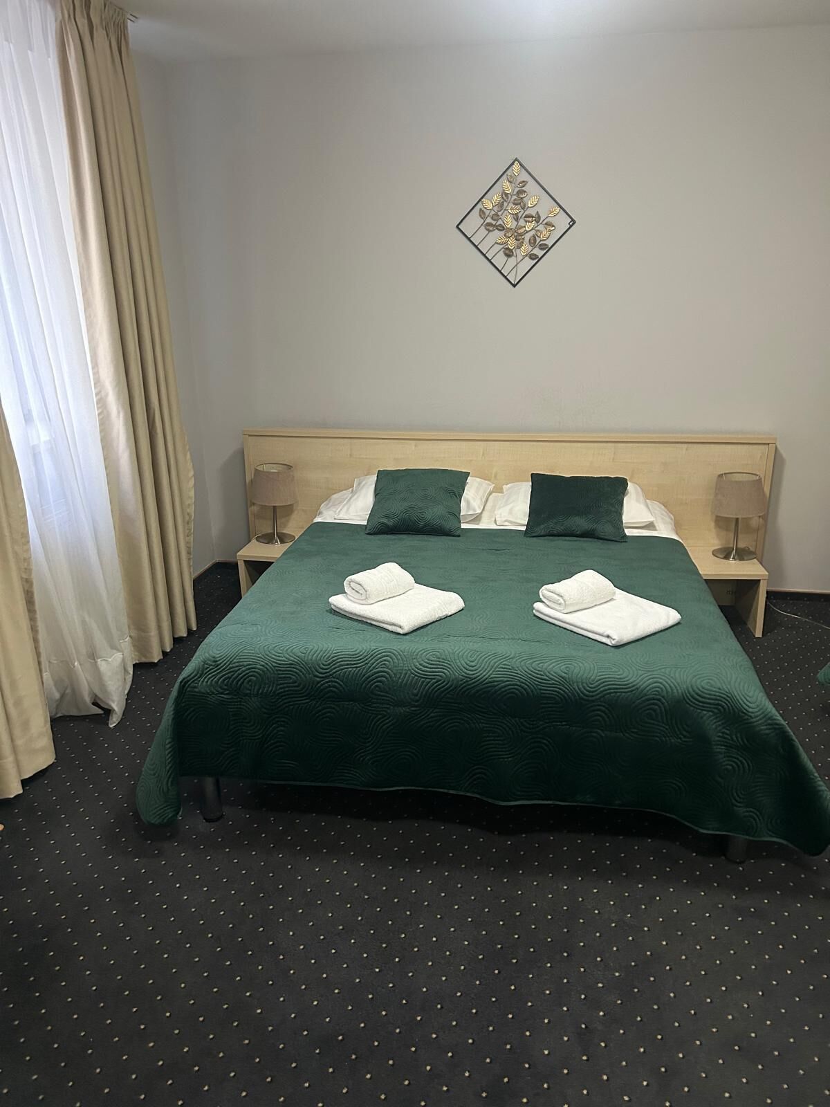 A neatly made bed with green bedding, two pillows, and two rolled towels. There is a wall decoration above the bed and a lamp on each side.