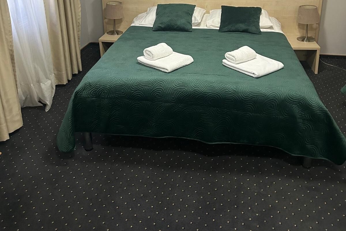 Een net opgemaakt bed met groen beddengoed, twee kussens en twee opgerolde handdoeken. Daarboven hangt een decoratie aan de muur en aan beide zijden staat een lamp.
