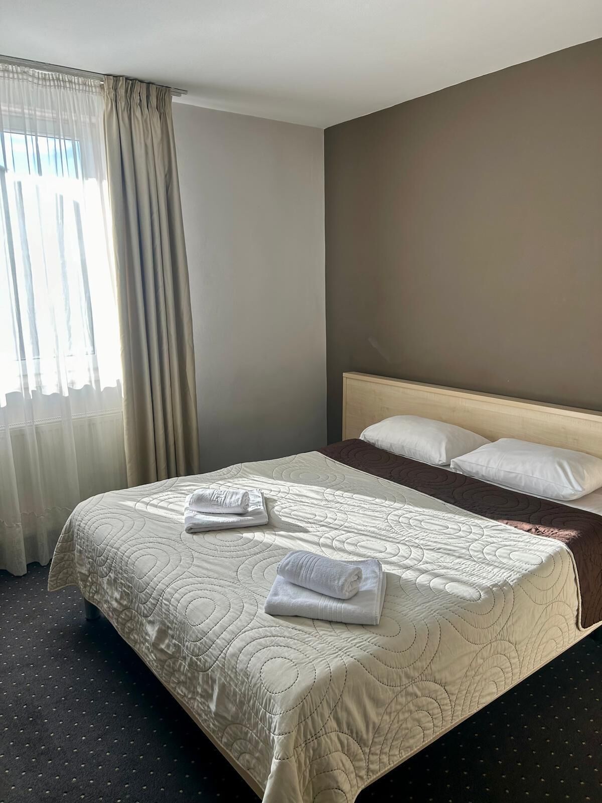 A neatly made bed with a beige quilt, two rolled towels, and a brown throw blanket.