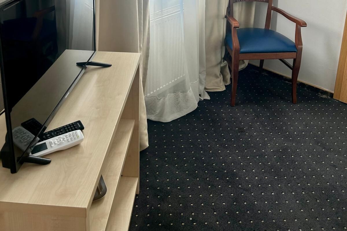 A hotel room with a TV on a wooden stand, a chair with a blue cushion, and a window with sheer curtains.