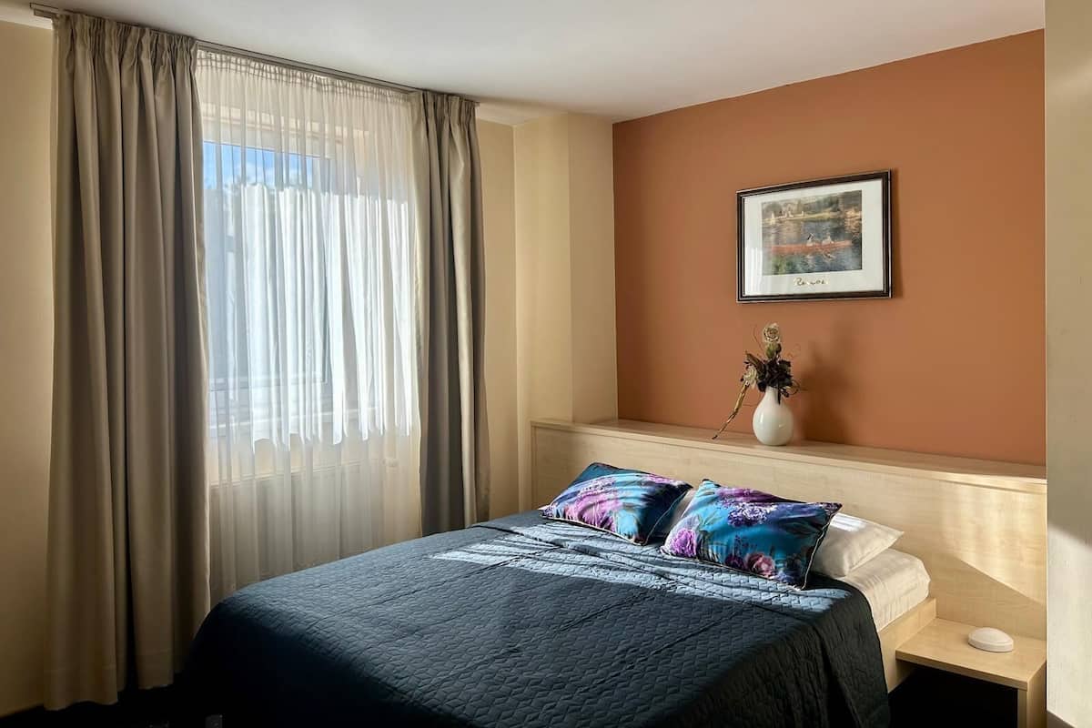 A hotel room with a bed, a nightstand, a vase with flowers, a framed picture on the wall, and sheer curtains.