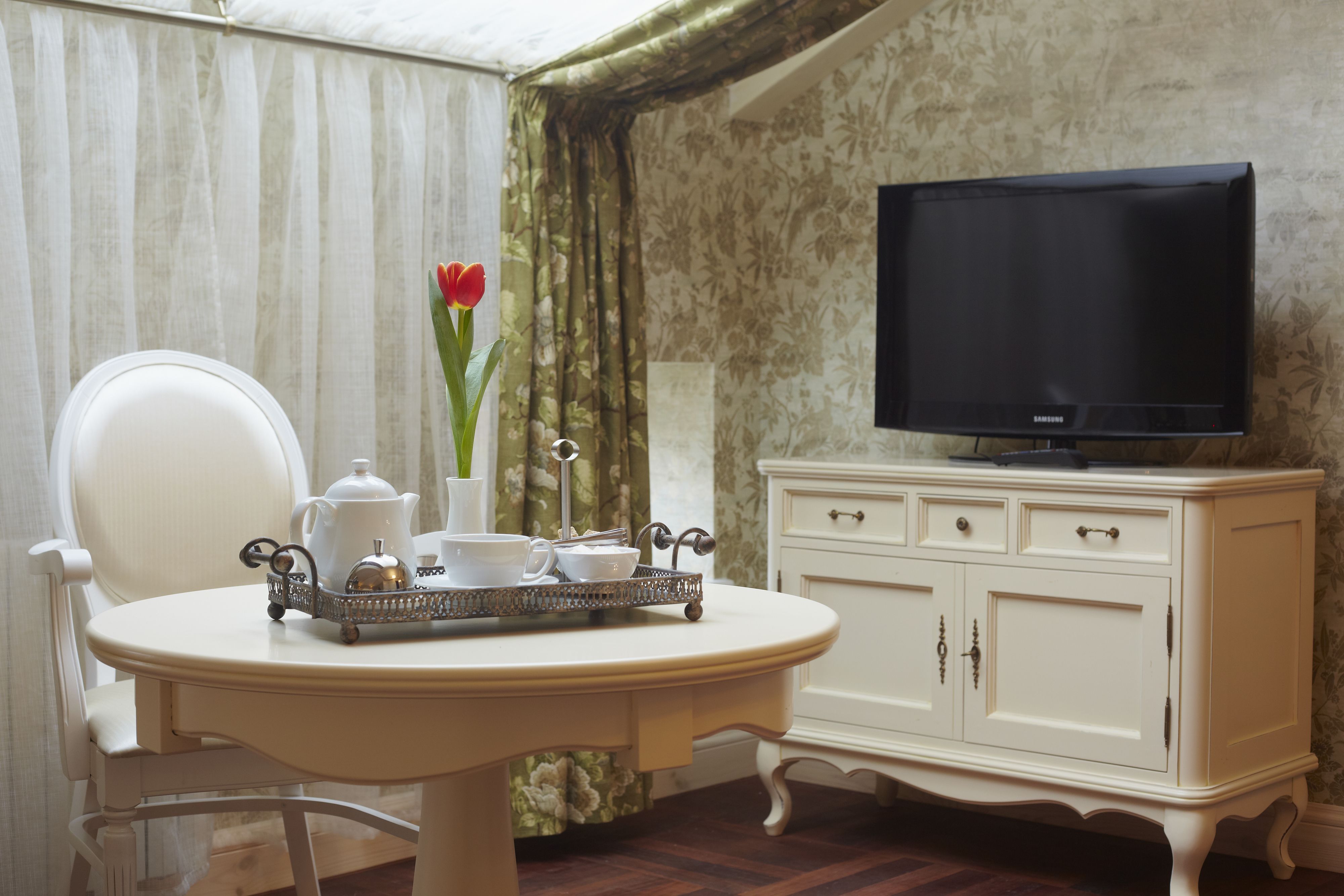 Una habitación con una mesa blanca dispuesta con una bandeja de tazas de té, una tetera y un florero con una flor roja. Hay una silla blanca, un televisor sobre un mueble blanco y una ventana con cortinas.