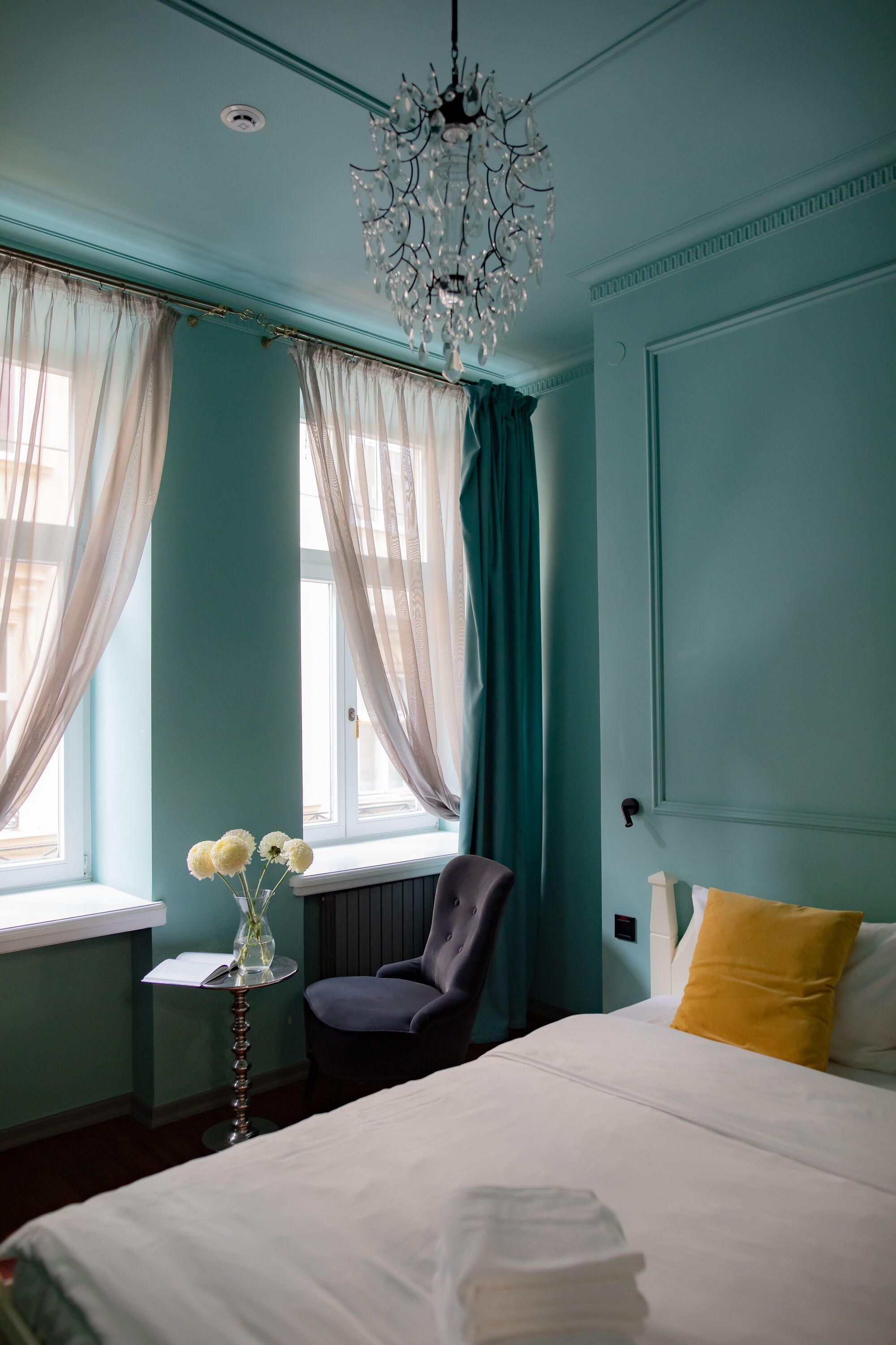Una habitación con paredes verde azulado, una cama blanca, una silla, una mesita con un jarrón de flores y una araña.