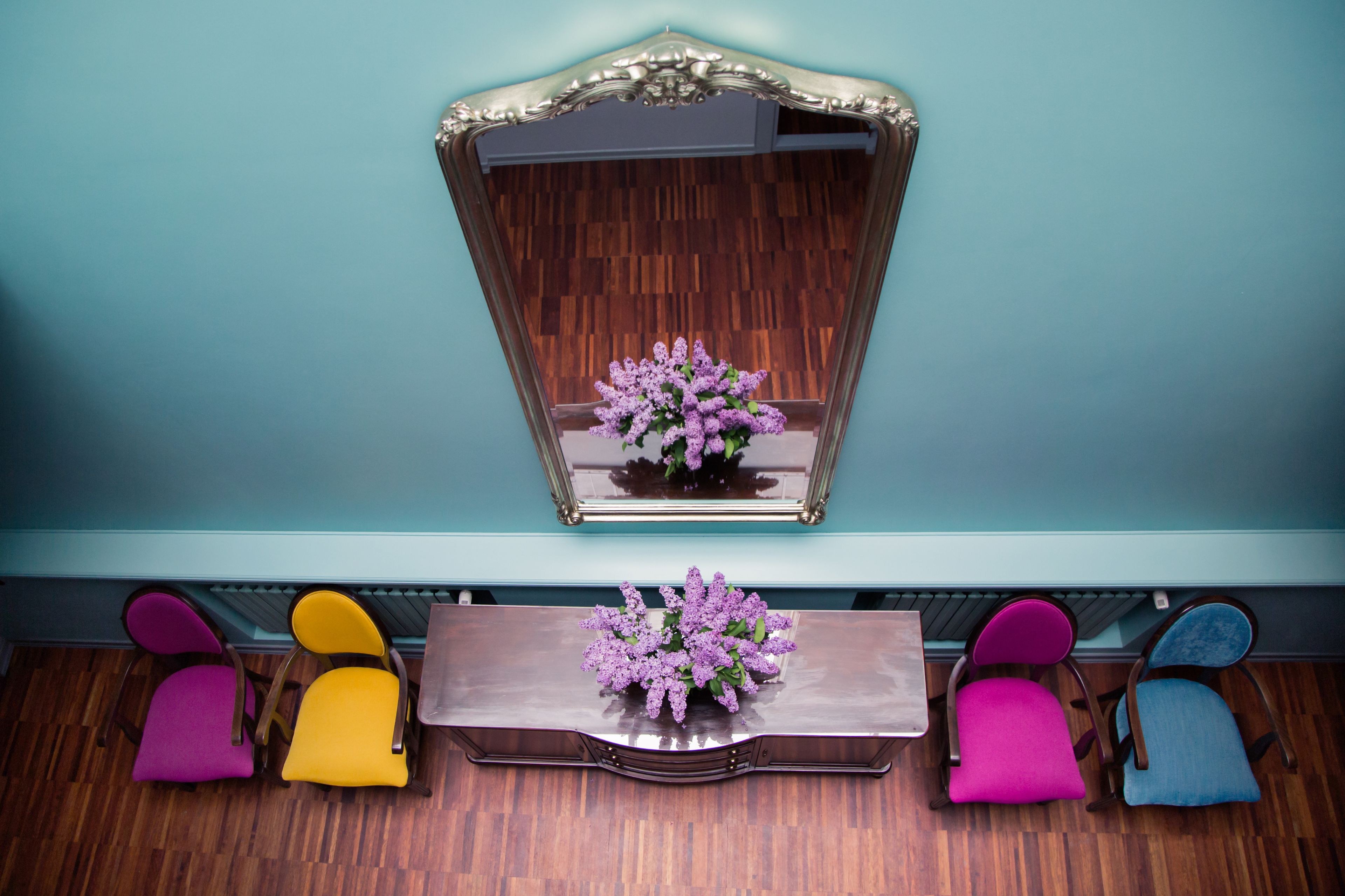 Una habitación con suelo de madera, pared color turquesa y un espejo sobre una mesa de madera con un arreglo floral púrpura. Hay cuatro sillas en tonos morado, amarillo, rosa y azul.