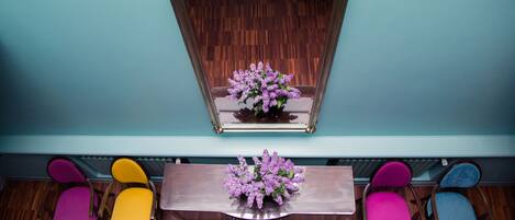 Una habitación con suelo de madera, pared color turquesa y un espejo sobre una mesa de madera con un arreglo floral púrpura. Hay cuatro sillas en tonos morado, amarillo, rosa y azul.